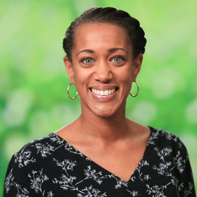 Smiling person with hoop earrings and a dark floral top, standing in front of a blurred green background.