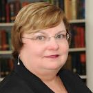 A person with short light brown hair, glasses, and earrings is wearing a dark blazer. They are in front of a blurred bookshelf background.