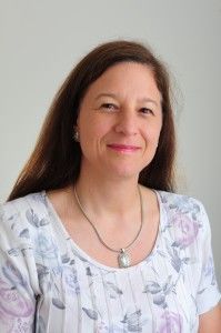 A woman with long brown hair is smiling, wearing a floral blouse and a necklace. She is posed against a plain, light-colored background.