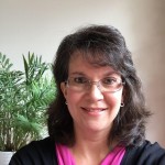 A person wearing glasses and a black top with a pink shirt smiles in front of a plant.