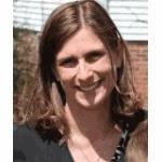 A person with shoulder-length brown hair is smiling outdoors, wearing a dark top. The background features a brick building and greenery, partially visible.