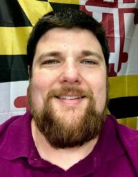 Man with a beard, wearing a purple shirt, smiles at the camera. The background features a Maryland flag.