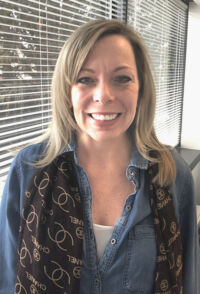 Woman smiling in front of window blinds, wearing a blue denim shirt and a patterned scarf.