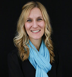 Smiling person with long, wavy blonde hair wearing a light blue scarf and black top against a dark background.